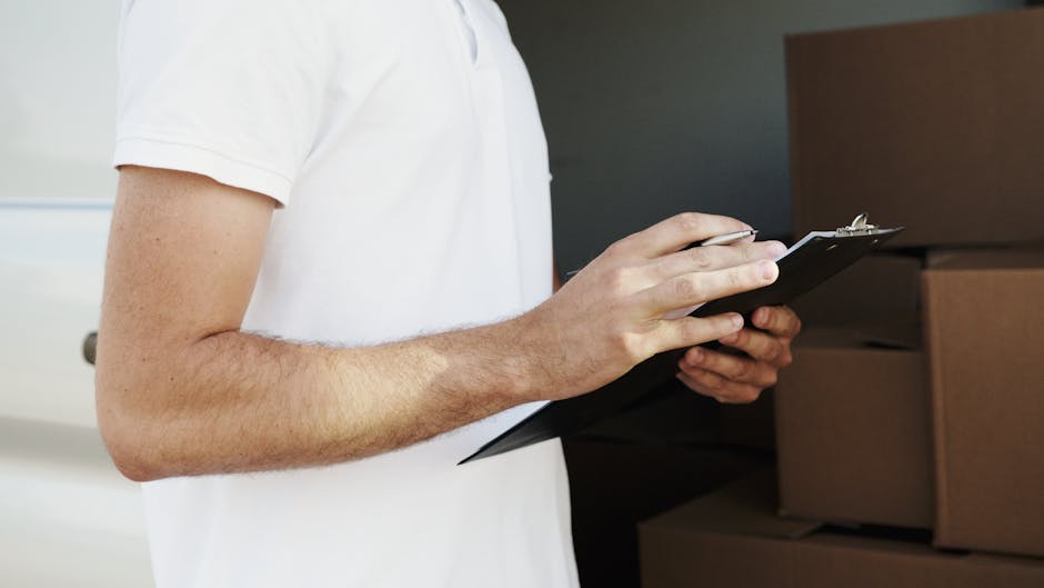 Close-up of a man wearing a white t-shirt, holding a black clipboard with a silver pen in his right hand, and inspecting or noting details during the home relocation process. In the background, there are several cardboard boxes stacked inside a room, some wrapped in plastic or fabric for protection. The scene suggests an organised packing and moving or furniture transport activity, with the man possibly overseeing the packing, loading, or inventory of belongings as part of a professional removals service. The environment is well-lit, and the focus is on the man's arm and hand, capturing the careful handling and documentation involved in house removals, aligning with the services provided by companies like Man with Van Harringay.