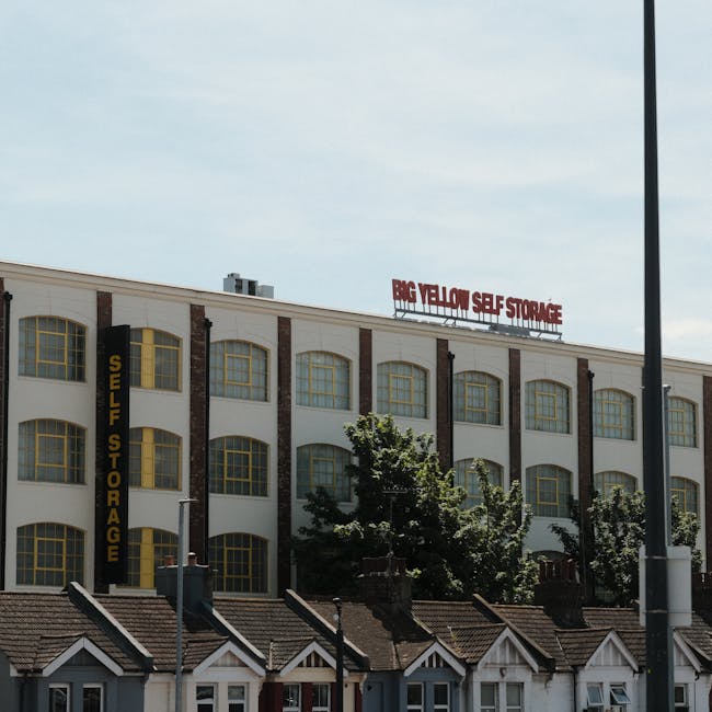 An exterior view of a multi-storey building with a sign reading 'BIG YELLOW SELF STORAGE' in red letters on the rooftop, and a vertical yellow 'SELF STORAGE' sign on the side, indicating a self-storage facility. The building has a white facade with large, evenly spaced windows featuring yellow frames, separated by vertical brown brick columns. In the foreground, several small residential houses with tiled roofs and white window frames are visible, along with a streetlight pole and trees partially obscuring the lower part of the building. The scene captures a bright, partly cloudy day, highlighting the location's role in home relocation and furniture storage services provided by companies like Man with Van Harringay.