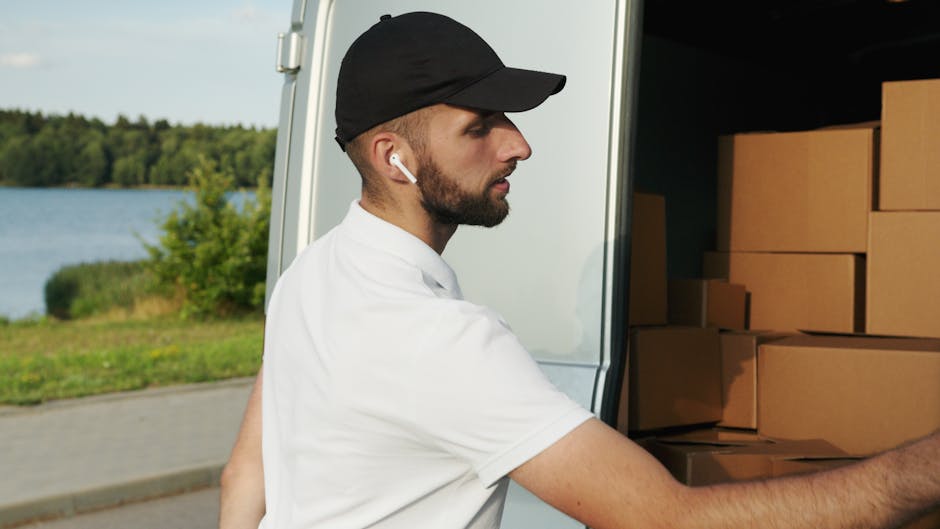 A male mover wearing a black cap, white T-shirt, and wireless earbud is seen outside near the back of a large moving van. Inside the van, several medium-sized cardboard boxes are stacked neatly, prepa
