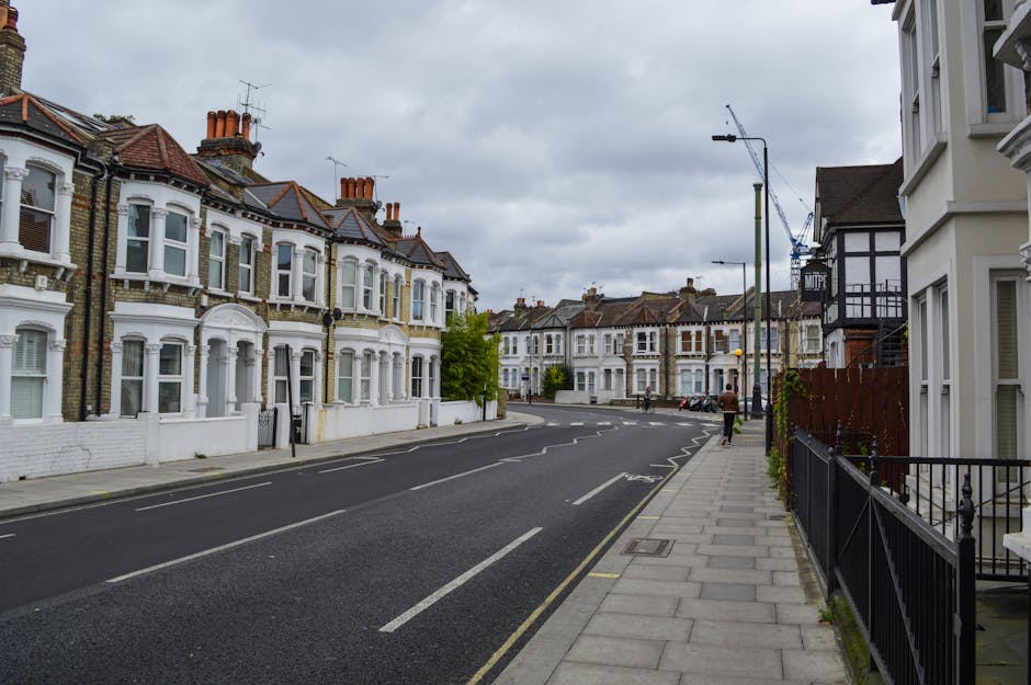 A quiet residential street with traditional Victorian terraced houses featuring white facades, bay windows, and red-tiled roofs, under an overcast sky. The pavement on the right side of the image has a black metal fence in front of a white house with a small garden area. Several cardboard boxes and packing materials are visible inside the doorway of one of the houses, indicating ongoing home relocation preparations. A few individuals can be seen in the distance, possibly involved in packing or moving activities. A man from Man with Van Harringay is loading or unloading belongings, using equipment such as a trolley to transport boxes along the sidewalk. A parked van is partially visible further along the street, suggesting the start of furniture transport and logistics associated with house removals. Street lamps, parked cars, and a crane in the background highlight the typical urban environment supporting efficient packing and moving operations.