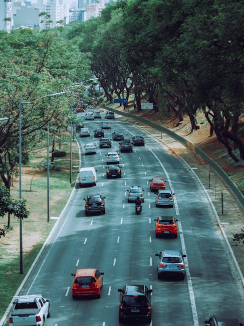 An aerial view of a busy residential street in Harringay with a row of parked cars along the curb and moving vehicles, including a red double-decker bus, driving on the road. The street is lined with trees, some of which are fully leafed, providing greenery to the area. On the left side, there are several brick and stucco buildings, including shops and residential properties, with pedestrians walking on the sidewalk. In the foreground, bicycles are parked near a small corner shop, and a few people are seen waiting at a crosswalk. The scene illustrates typical urban activity during daytime, with a focus on residential and commercial environments suitable for coordinated house removals or furniture transport that a company like Man with Van Harringay might facilitate during a home relocation, supporting efficient packing and moving logistics.