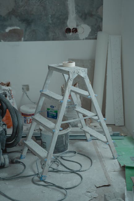 A white, four-step folding wooden ladder positioned in a room under renovation with exposed wall patches and partially finished surfaces. To the left of the ladder, a black vacuum cleaner is visible, alongside a container of paint or filler and some electrical cords lying on the floor. Behind the ladder, there are several white wall panels or boards leaning against the wall, with construction materials and tools scattered around, indicating an ongoing home improvement or packing process. This setting illustrates a typical environment for house removals or furniture transport preparations, with the ladder likely used for accessing higher areas during packing or reassembly, consistent with the context of professional removals services like those provided by Man with Van Harringay.