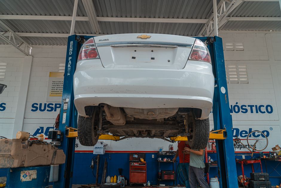 A white Chevrolet sedan is elevated on a blue hydraulic lift inside an automotive workshop. The car is suspended with its underside visible, showing the exhaust system, suspension components, and tires on either side. The workshop has white brick walls with blue lettering spelling out words related to automotive diagnostics and maintenance. There are various tools, equipment, and containers on workbenches and shelves along the background. Two technicians, partially visible, are working beneath the car; one holds a tool while the other observes. Overhead lighting illuminates the workspace, highlighting the car's undercarriage and surrounding tools, reflecting a professional environment for vehicle repair and inspection, aligned with the proper procedures used in home relocation vehicle transport and maintenance services by Man with Van Harringay.