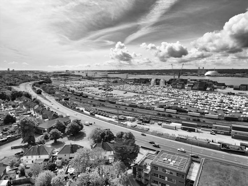 A wide aerial black and white photograph showing a dense industrial port area adjacent to a river, with numerous storage containers, cranes, and warehouses visible in the distance. In the foreground, residential houses with pitched roofs and surrounding trees are seen along a curving road, leading towards the port. The scene captures a busy environment associated with logistics and freight transport, illustrating the scale of goods movement that might be involved in house removals or furniture transport services provided by Man with Van Harringay. The sky is partly cloudy with scattered clouds and streaks of sunlight breaking through, providing natural lighting over the entire landscape.