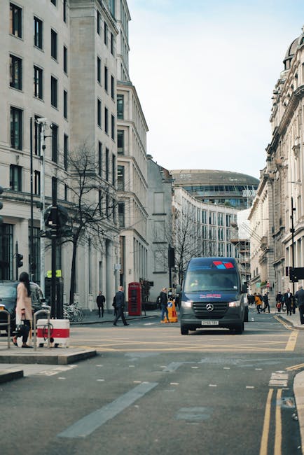 A view of a busy city street with tall, white and beige multi-storey buildings on both sides. In the center, a blue van with red markings on the windshield is parked, likely being used for a home relocation or furniture transport. Several pedestrians are walking along the sidewalks, some crossing the street, while others stand near the curb. The pavement shows safety cones and a red British telephone booth is visible on the left side of the street. The scene is during daylight hours with clear skies, and the environment suggests an urban area suitable for carrying out house removals. The image highlights the logistical aspects of moving, with the van positioned for loading or unloading furniture or packed boxes, illustrating typical elements encountered during professional removals in Haringey, as referenced on the 'Turnpike Lane man with van access guide' page for Man with Van Harringay.